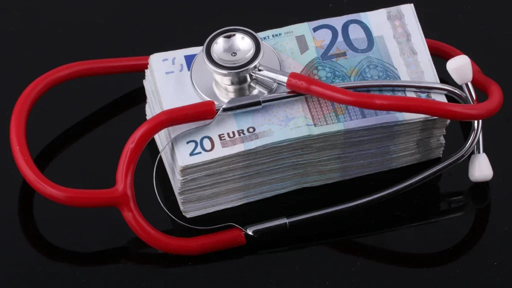 Medicīnas stetoskops novietots uz eiro banknošu kaudzes, uzsverot finanšu kontroli veselības aprūpes nozarē.