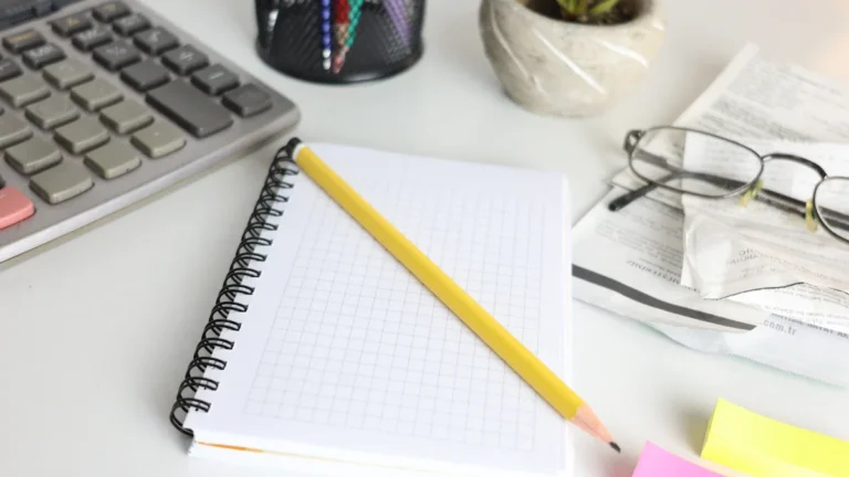 Rakstāmgalds ar kalkulatoru, piezīmju grāmatiņu un brillēm, simbolizējot nodokļu aprēķinus un darba plānošanu.
