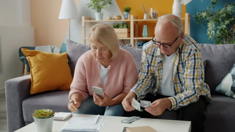 Senioru pāris mājās sēž pie galda, skaita skaidru naudu un pārskata finanšu dokumentus.