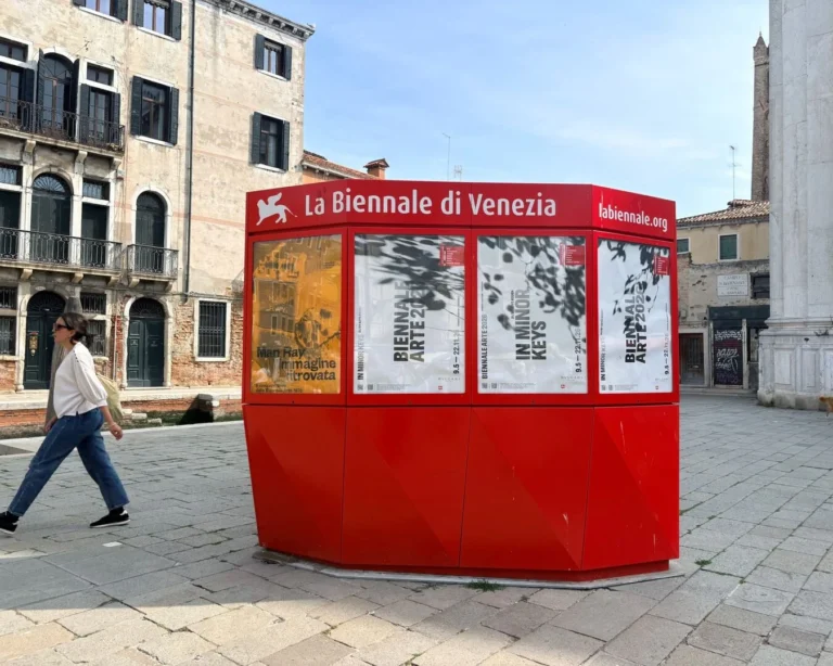 Sarkans Venēcijas biennāles informācijas stends ar plakātiem pilsētas laukumā, fonā vēsturiskas ēkas.