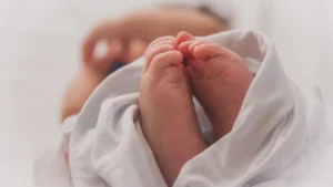 Jaundzimuša bērna pēdas baltā segā, attēlotas tuvplānā uz neitrāla, gaiša fona.