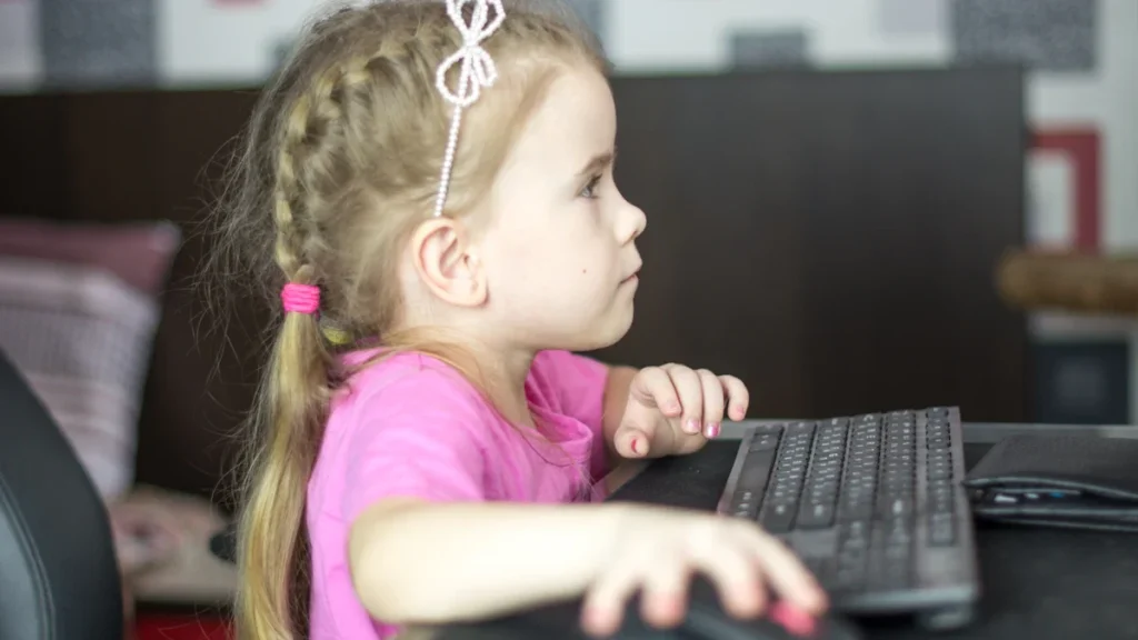 Maza meitene sēž pie datora tastatūras un peles, koncentrēti skatoties ekrānā mājas vidē.