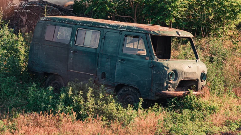 Vecs, sarūsējis zaļas krāsas mikroautobuss pamests pļavā, simbolizējot nolietotus transportlīdzekļus.