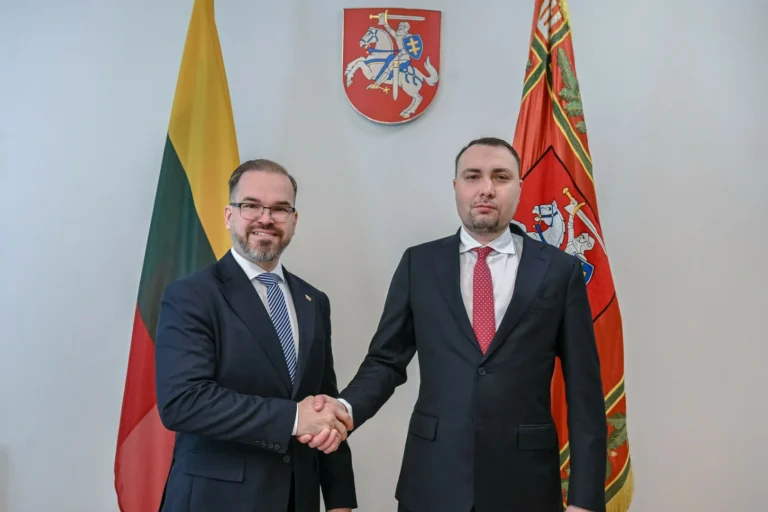 Divi vīrieši uzvalkos sarokojas telpā pie Lietuvas karogiem un valsts ģerboņa pie sienas.