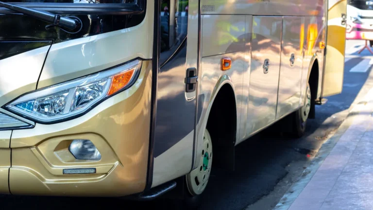Tuvplāns no starppilsētu autobusa priekšpuses un sāna uz asfalta seguma pilsētvidē saulainā dienā.