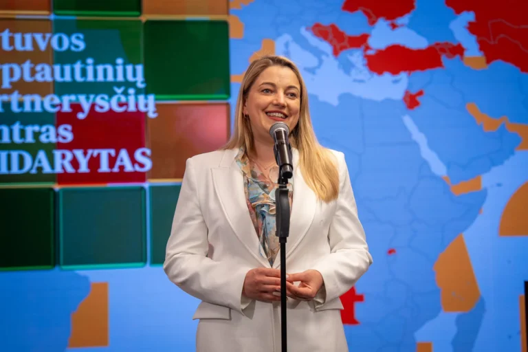 Smaidīga sieviete baltā žaketē runā mikrofonā uz krāsaina pasaules kartes un teksta fona.