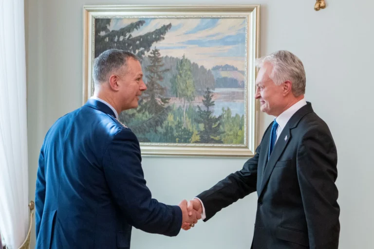 Militārpersona zilā uniformā un vīrietis tumšā uzvalkā sarokojas pie gleznas ar dabas ainavu.
