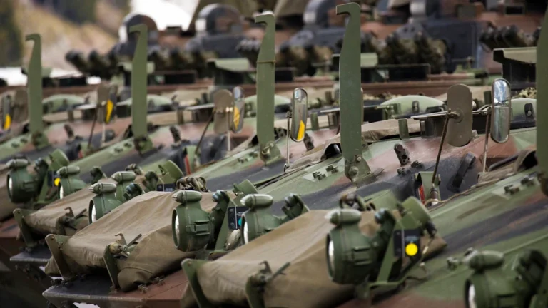 Rinda ar moderniem kamuflāžas krāsas bruņutransportieriem, kas izvietoti ar militāru precizitāti.