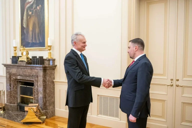 Divi vīrieši uzvalkos spiež rokas gaišā telpā pie marmora kamīna un senatnīgas gleznas.