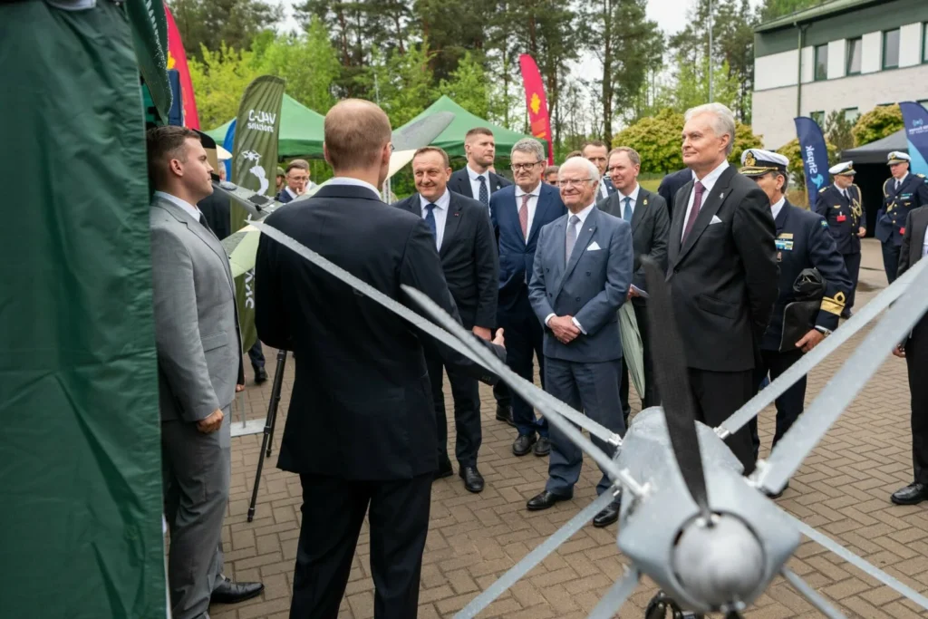 Vīriešu grupa uzvalkos stāv ārā pie pelēka bezpilota gaisa kuģa ar asiem propelleriem.