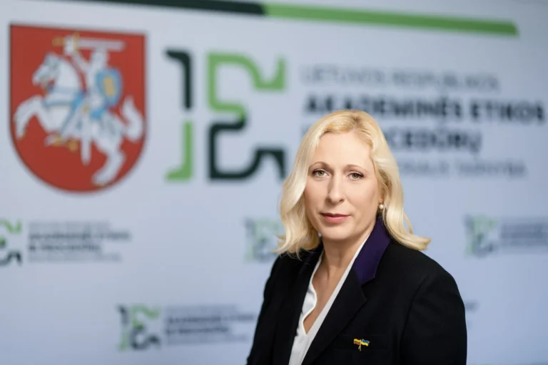 Sieviete ar blondiem matiem melnā žaketē uz fona ar Lietuvas ģerboni un organizācijas logotipu.