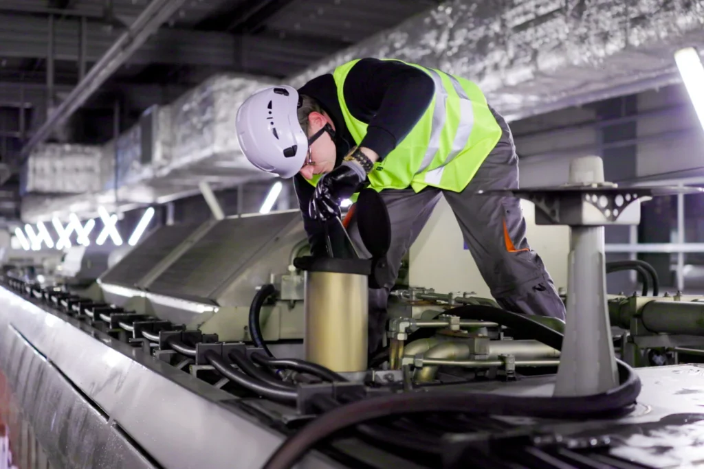 Darbinieks baltā ķiverē un dzeltenā drošības vestē veic industriālu iekārtu apkopi tehniskajā telpā.