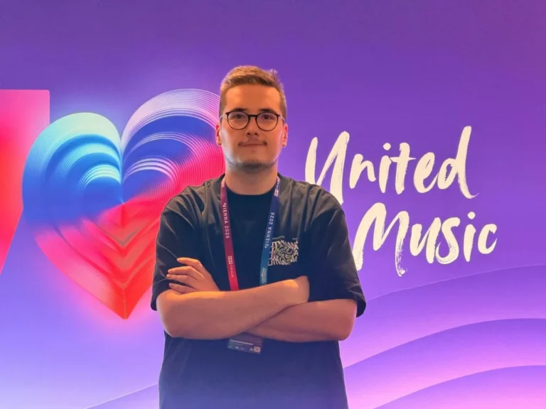 Jauns vīrietis brillēs ar sakrustotām rokām uz violeta fona, kurā redzama sirds un teksts "United Music".
