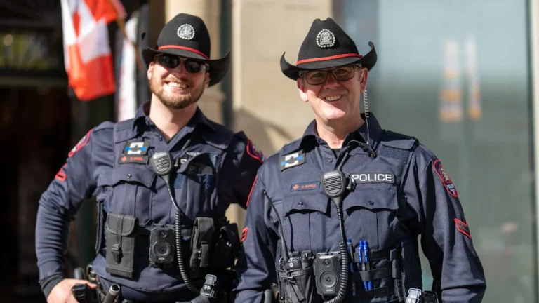 Divi pieredzējuši policijas darbinieki formastērpos smaidot stāv blakus, simbolizējot ilggadēju un godprātīgu dienestu.