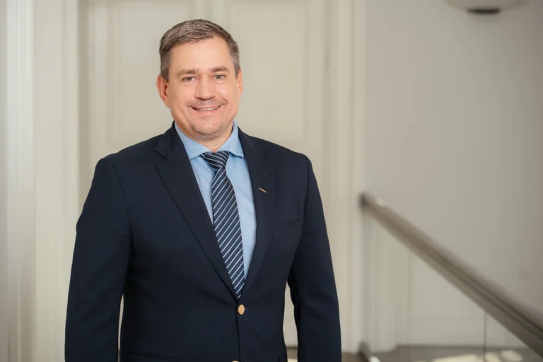 Smaidīgs vīrietis tumši zilā žaketē, gaiši zilā kreklā un svītrainā kaklasaitē uz gaiša telpas fona.