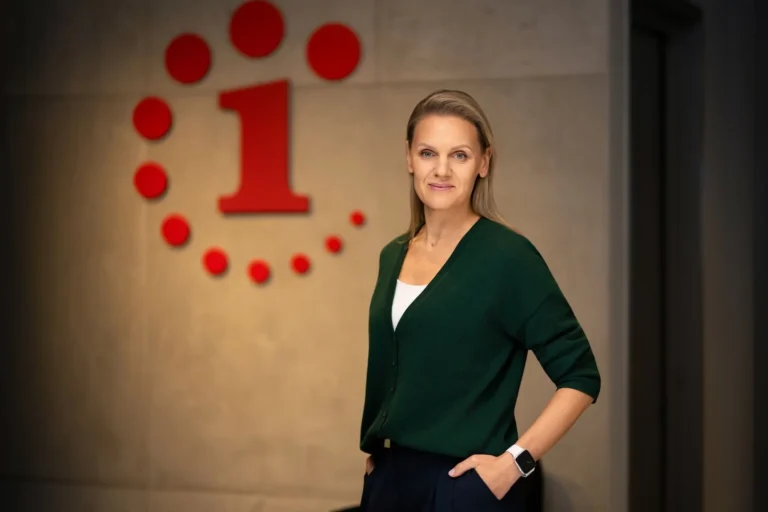 Gaišmataina sieviete zaļā jaciņā stāv pie sienas ar Latvijas Radio 1 sarkano logotipu.