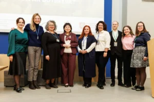 Deviņu smaidīgu cilvēku grupa stāv telpā pie prezentācijas ekrāna oficiālā pasākumā.
