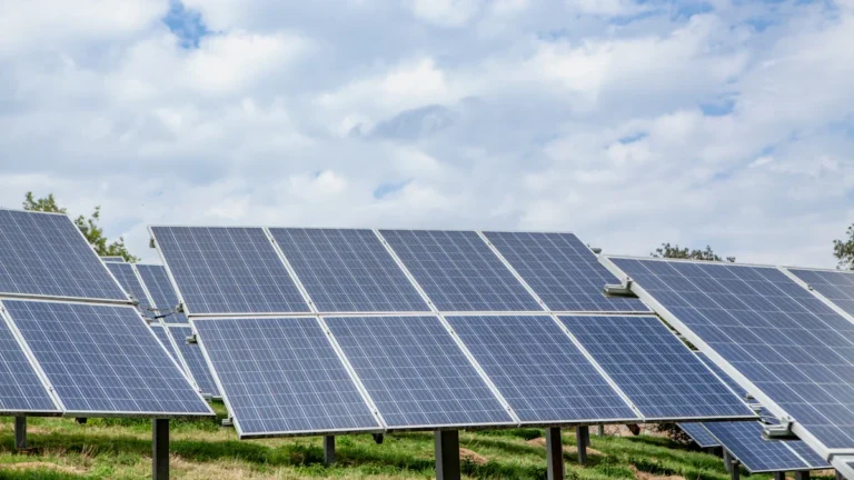 Modernu saules paneļu rindas zem mākoņainām debesīm, attēlojot atjaunojamās enerģijas infrastruktūru lielā mērogā.