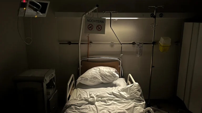 Moderna slimnīcas gulta un medicīniskais monitors pie sienas gaišā, tukšā palātā.