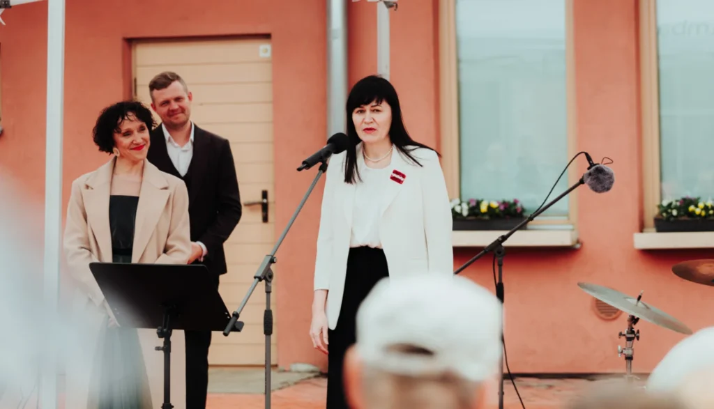 Sieviete baltā žaketē ar Latvijas karoga piespraudi uzstājas pie mikrofona pasākumā pie ēkas.