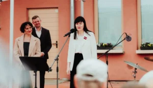 Sieviete baltā žaketē ar Latvijas karoga piespraudi uzstājas pie mikrofona pasākumā pie ēkas.