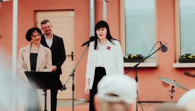 Sieviete baltā žaketē ar Latvijas karoga piespraudi uzstājas pie mikrofona pasākumā pie ēkas.