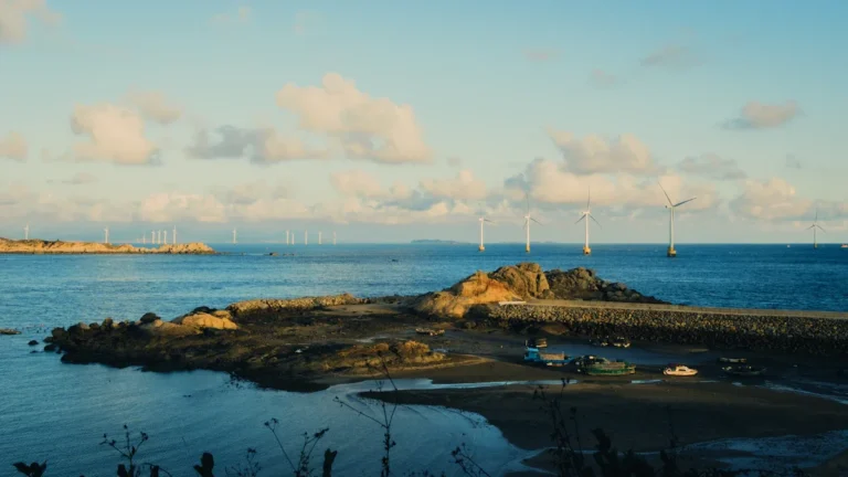 Skats uz jūras piekrasti ar akmens molu un vēja turbīnām horizontā, simbolizējot piekrastes attīstību.
