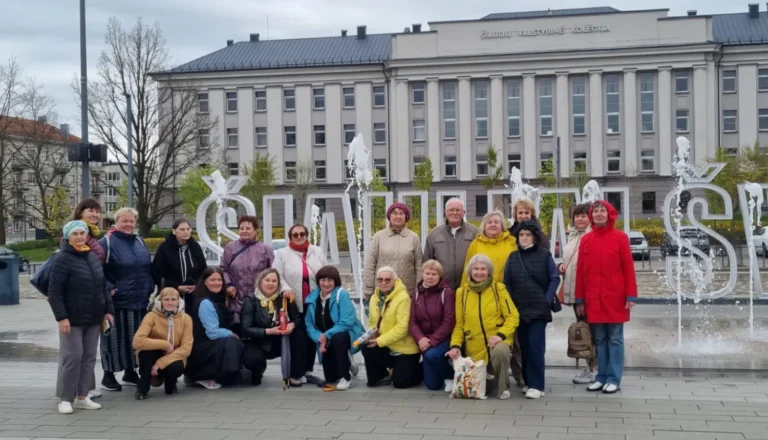 Cilvēku grupa pozē pie strūklakas un liela uzraksta Šauļos pie Šauļu valsts koledžas ēkas.