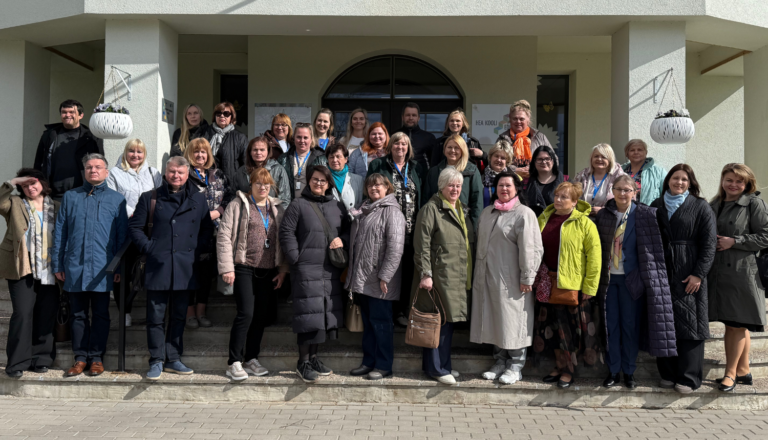 Liela pieaugušo grupa mēteļos stāv uz kāpnēm pie gaišas ēkas ieejas kopbildē.