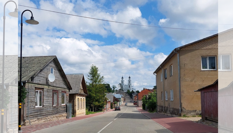 Asfalta iela ar koka māju kreisajā pusē un baltu divtorņu baznīcu tālumā zem mākoņainām debesīm.