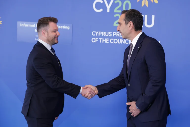 Divi vīrieši tumšos uzvalkos sarokojas uz zila fona ar Kipras prezidentūras tekstu un logo.