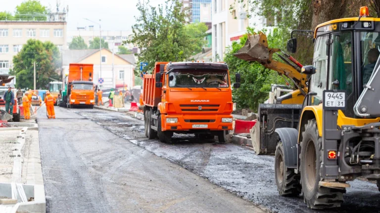 Ceļu būves tehnika un oranžas kravas automašīnas veic asfalta seguma remontdarbus uz pilsētas ielas.