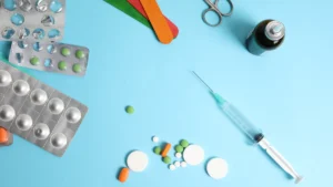 Medicīniskie piederumi, tabletes iepakojumos un šprice uz zila fona, simbolizējot veselības aprūpes pakalpojumus.