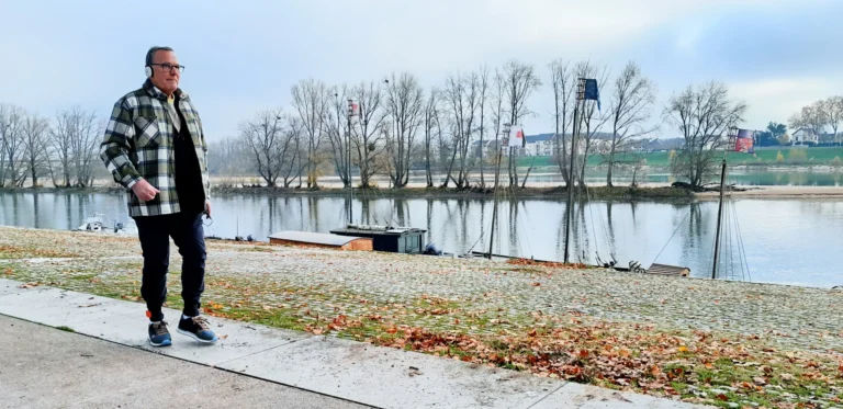 Vīrietis rūtainā jakā un austiņās soļo pa bruģētu krastmalu gar upi, kurā pietauvotas laivas.