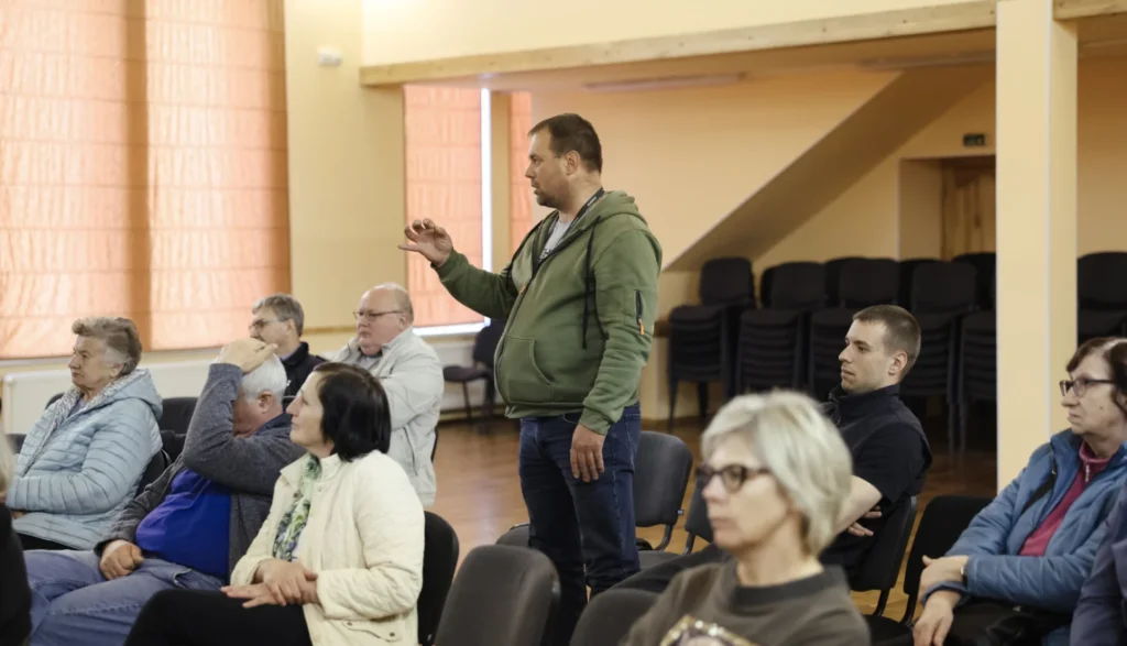 Vīrietis zaļā džemperī stāv un runā auditorijas priekšā sanāksmju telpā, kurā sēž vairāki cilvēki.