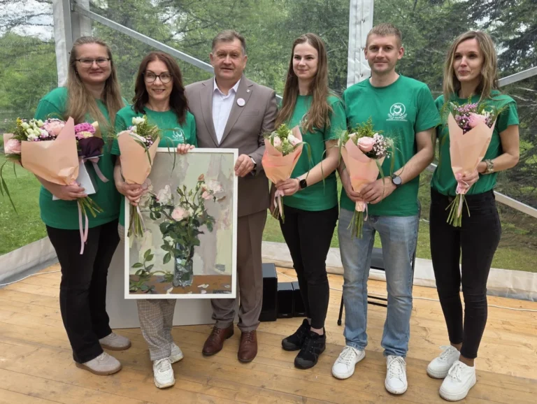 Seši cilvēki pozē ar ziedu pušķiem un rožu gleznu gaišā teltī uz koka grīdas.
