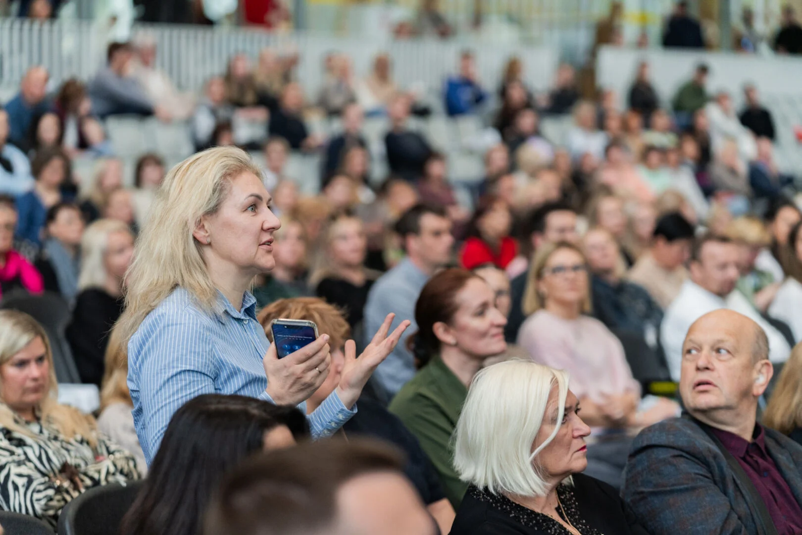 Gaišmataina sieviete zilā strīpainā kreklā stāv un žestikulē, uzrunājot auditoriju pilnā konferenču zālē.