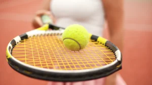 Tenisa bumbiņa uz raketes stīgām uz sarkana māla laukuma fona, simbolizējot Roland Garros atmosfēru.