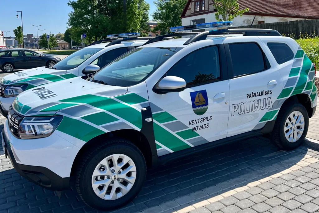 Divas baltas Ventspils novada pašvaldības policijas automašīnas ar zaļām svītrām un zilām bākugunīm.