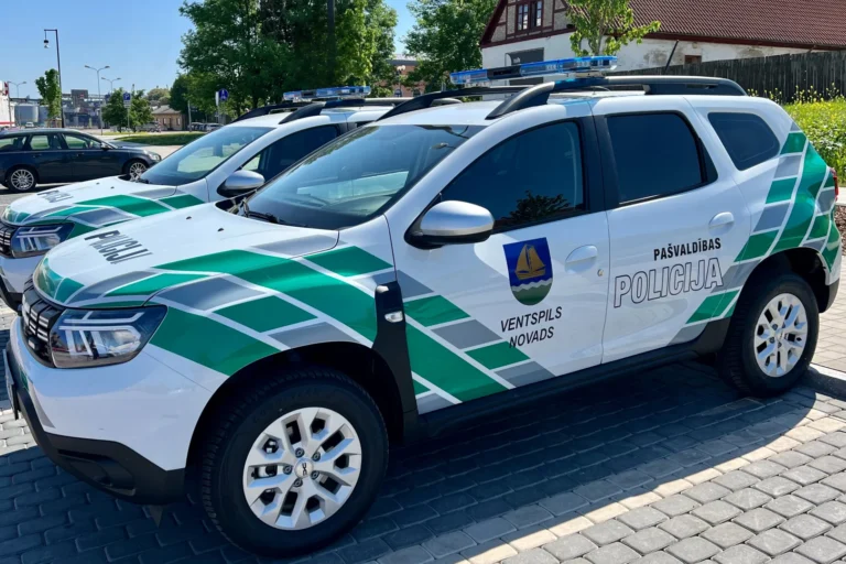Divas baltas Ventspils novada pašvaldības policijas automašīnas ar zaļām svītrām un zilām bākugunīm.