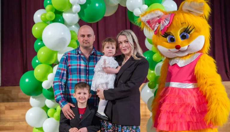 Ģimene ar bērniem pozē pie balonu arkas kopā ar lielu, dzeltenu lapsas kostīma tēlu.