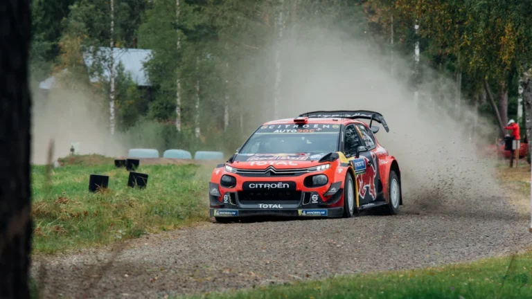 Sarkana un melna rallija automašīna lielā ātrumā traucas pa grants ceļu mežainā apvidū.