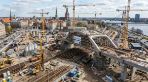 Rail Baltica būvlaukums Rīgā ar dzelteniem celtņiem, tilta konstrukcijām un Vecrīgas panorāmu fonā.