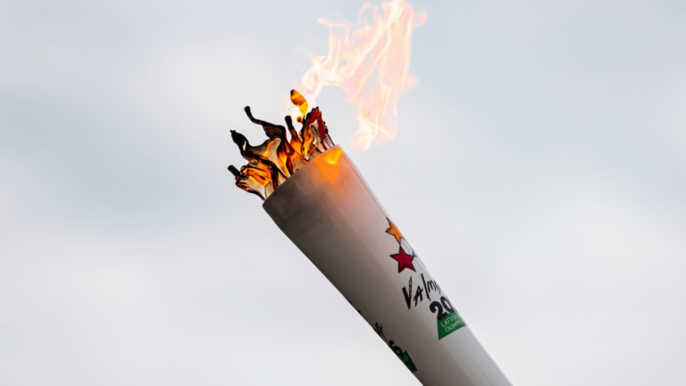 Degoša balta lāpa ar krāsainu zvaigžņu logotipu un tekstu uz gaišu, dūmakainu debesu fona.