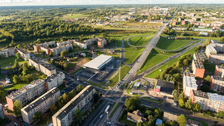 Pilsētas panorāma no putna lidojuma ar satiksmes pārvadu, daudzdzīvokļu ēkām un plašu autostāvvietu.