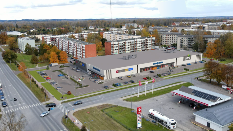 Aerofotoattēls ar modernu tirdzniecības centru, plašu autostāvvietu un apkārtējām daudzdzīvokļu mājām rudenī.