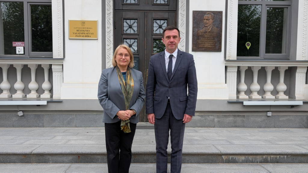 Sieviete un vīrietis lietišķos uzvalkos stāv pie Rēzeknes valstspilsētas pašvaldības ēkas ieejas.