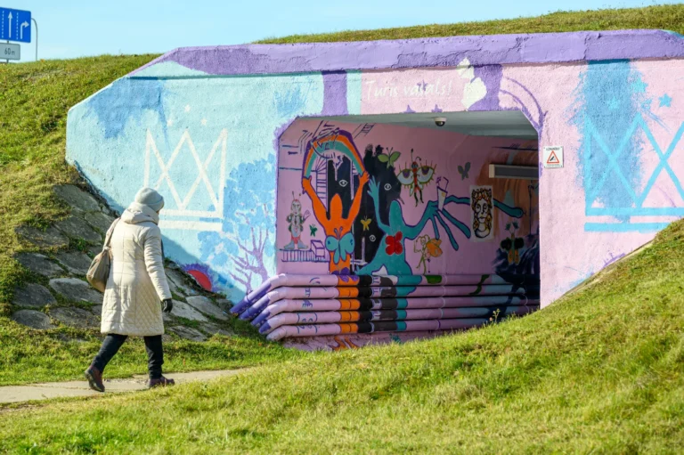 Gājēja gaišā ziemas mētelī iet garām koši apgleznotai tuneļa ieejai ar dažādiem grafiti zīmējumiem.