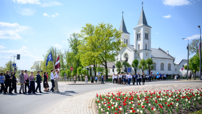 Cilvēku grupa ar Latvijas karogu gājienā gar baltu divtorņu baznīcu pie ziedošas tulpju dobes.