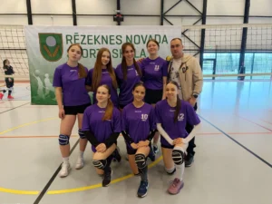 Septiņas volejbolistes violetos tērpos un viņu treneris pozē pie Rēzeknes novada sporta skolas banera.